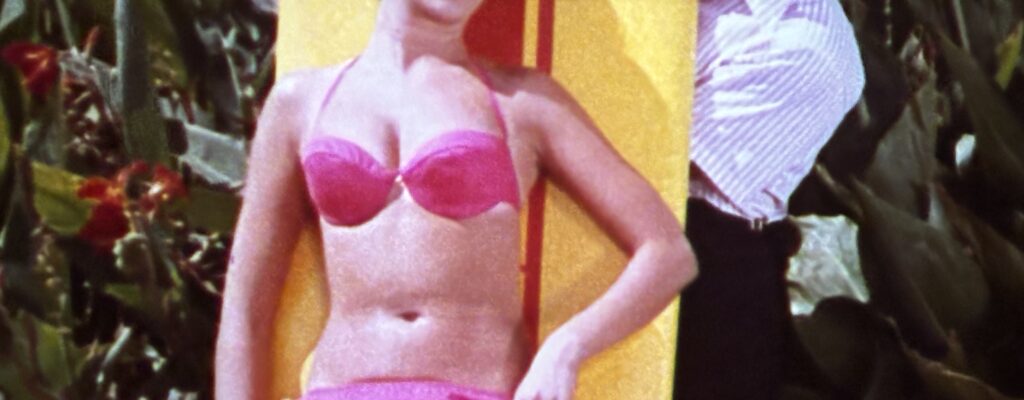 A woman dressed in a 1960s-era bikini stands in front of a bright yellow surfboard. A man peeks out from behind the surfboard and looks at the woman.